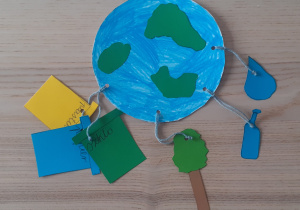 Do wyciętej z papieru kuli ziemskiej przyczepione są papierowe przywieszki: zielone drzewko, niebieska kropla, zielony, niebieski i żółty trapez symbolizujący kontenery na segregację śmieci.